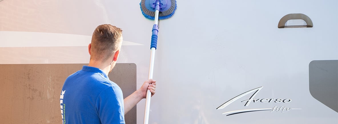 Tout beau tout propre : comment préparer ma caravane pour la nouvelle saison ? Personne en t-shirt bleu nettoyant un camping-car Averso avec une brosse télescopique bleue, vue de dos