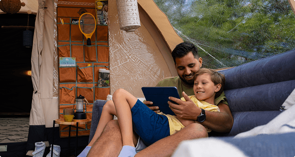 père et fils sur un canapé gonflable Père et fils se détendant dans une tente de glamping confortable, regardant une tablette ensemble, avec des accessoires de camping organisés en arrière-plan