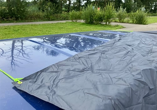 Rabatt de connexion de la tente pour van installé sur le toit de la voiture. Rabatt de connexion d’une tente pour van avec hauban, posé sur une surface bleu foncé dans un environnement verdoyant avec des arbustes et des arbres.
