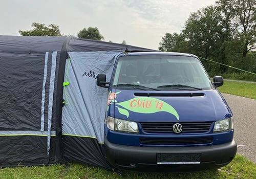 Auvent de camping pour Volkswagen T4 en plein air Fourgon Volkswagen bleu avec auvent de camping attaché sur un terrain herbeux par temps clair, idéal pour les voyages en plein air