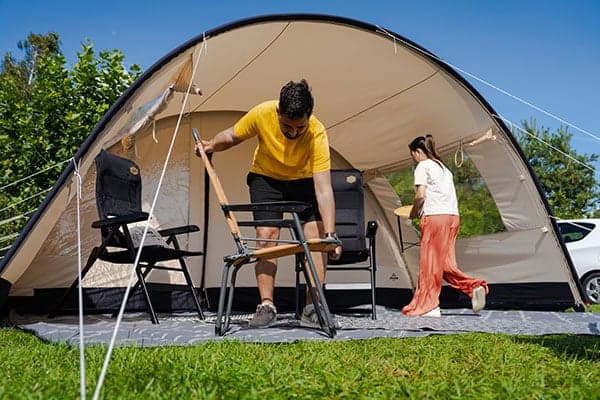 Homme dépliant une chaise devant une tente Tente dôme spacieuse avec auvent sur une pelouse, deux personnes installent des chaises et une table de camping sous un ciel bleu ensoleillé